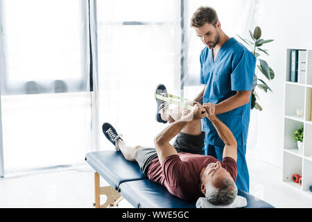 Beau médecin barbu tenant de la jambe du patient mature l'entraînement avec bandes élastiques Banque D'Images