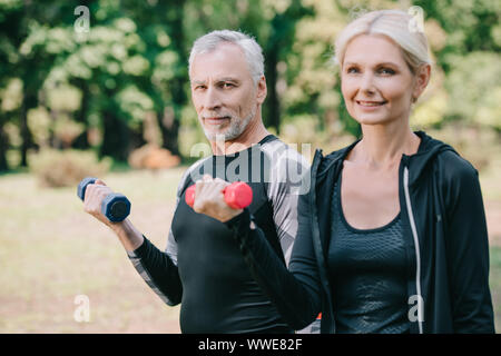 Smiling mature sportif et de la sportive à la caméra à alors que la formation avec barbells in park Banque D'Images