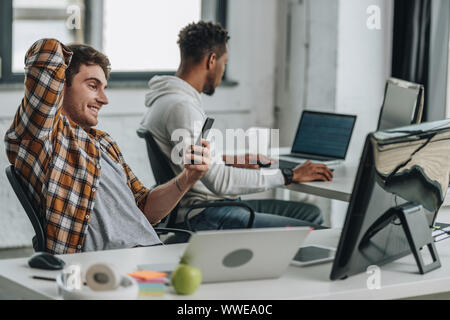 Programmeur gaie using smartphone tout en étant assis sur le lieu de travail près de african american collègue Banque D'Images