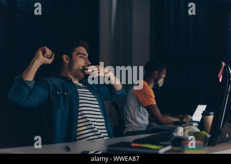 Fatigué de bâillements programmeur tout en restant assis près de african american collègue et nuit à l'office de tourisme Banque D'Images