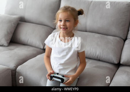 Fille blonde drôle sans une dent joue à des jeux vidéo dans un t-shirt blanc à la maison Banque D'Images