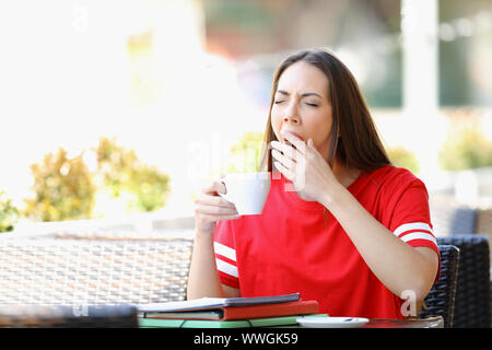 Fatigué student yawning dans un bar tenant une tasse de café assis dans un bar terrasse Banque D'Images