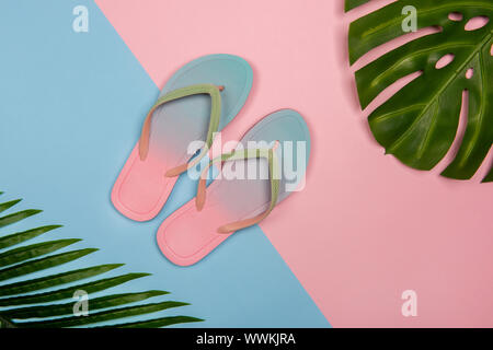 Les tongs plage élégante sur fond rose et bleu pastel avec monstera et plam feuilles, vue d'en haut. Concept minimaliste de l'été avec l'exemplaire de l'espace. Banque D'Images