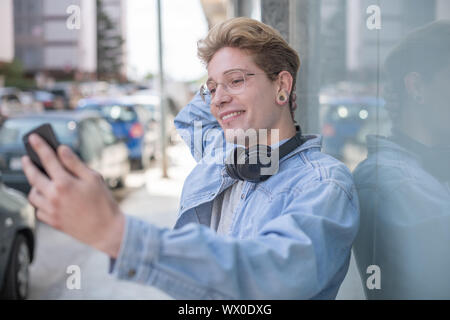 Portrait d'un garçon blond avec des lunettes et des écouteurs de prendre une photo de lui-même avec son smartphone dans la rue. Concept de jeunesse technologique. Banque D'Images