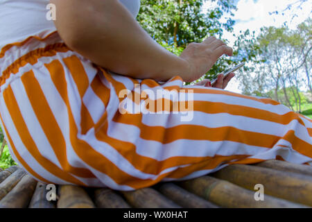 Femme assise sur le banc en bambou au jardin tropical et holding mobile phone. Galerie d'images haute résolution. Banque D'Images