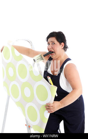 Exaspéré woman hanging wallpaper avec un pinceau plein de pâte colle serrée dans sa bouche comme elle tient le rouleau de papier à deux mains Banque D'Images