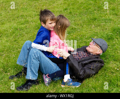 Deux enfants (frère et sœur - 7 et 5 ans) assis sur leur grand-père couché dans l'herbe Banque D'Images