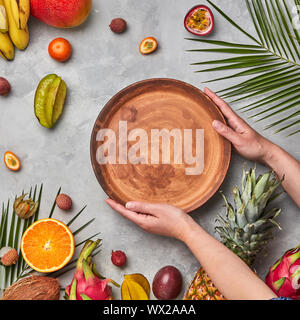 Mains d'une femme tenant une plaque vide brun autour d'un autre venu de fruits tropicaux sur un fond de béton gris avec copie savs Banque D'Images