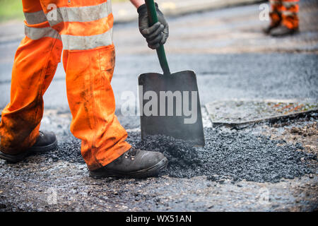 L'homme en pantalon de travail orange avec une pelle répandre de goudron et de gravier sur une route d'être refait surface Banque D'Images