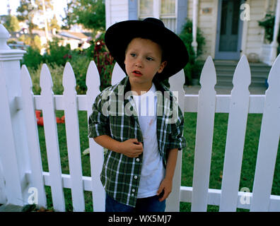 Garçon portant un chapeau noir debout à côté d'une clôture blanche à l'extérieur de sa maison. Banque D'Images