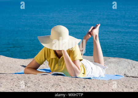 Womand de lire un livre en position couchée sur un rocher au bord de la mer Banque D'Images