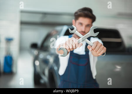 Beau jeune mécanicien en uniforme tenant deux clés est traversé en voiture centre de service. Banque D'Images