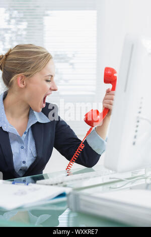 Jeune femme crier en direction du récepteur téléphonique rouge at desk in office Banque D'Images