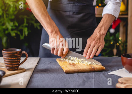 Un male chef dans un tablier noir prépare un plat de Noël par tranchage de l'ail sur une planche à découper. La cuisine joyeuse fête de Noël et Nouvel An le tableau Banque D'Images