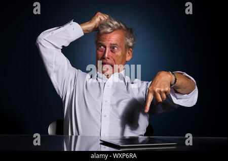 Émotionnelle très frustré senior business man pointing at his tablet computer allongé sur le bureau tandis que de déchirer ses cheveux avec sa main Banque D'Images