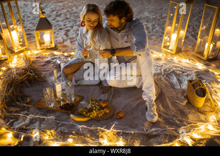 Couple ayant un dîner romantique, serrant et tintement des verres de vin à la place joliment décorées avec lumineux lumières différentes la plage de sable au crépuscule Banque D'Images