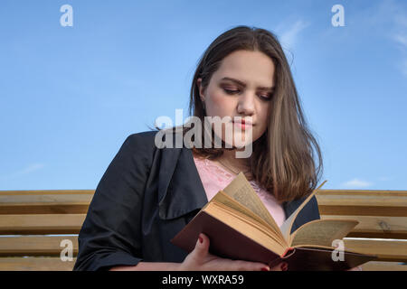 Brunette girl reading book sur un banc dans la rue. Fille en veste noire avec des cheveux longs avec réserve en journée d'été. Modèle de taille plus Banque D'Images