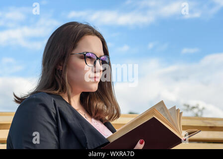 Brunette girl reading book sur un banc dans la rue. Fille en veste noire avec des cheveux longs avec réserve en journée d'été. Modèle de taille plus Banque D'Images