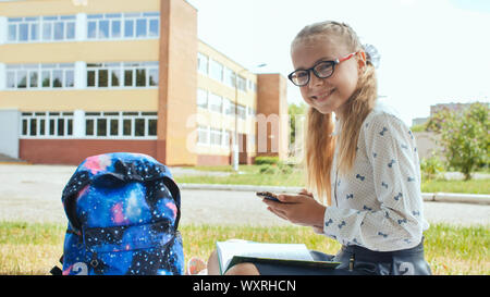 Une période de 11 ans, lycéenne est assis sur l'herbe avec un livre et cherche dans le téléphone. Banque D'Images