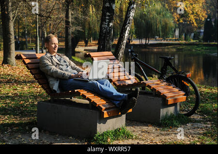 L'homme sur une chaise longue près du lac travaille sur un ordinateur portable. Un vélo est à proximité. Banque D'Images