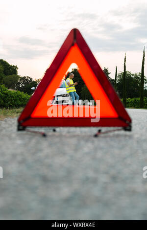 Signe d'arrêt d'urgence rouge et jeune homme en attente l'assistance voiture avec voiture cassée sur la route Banque D'Images