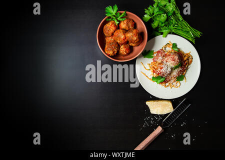 Un gros plan d'une assiette de spaghetti et de boulettes de viande rustiques avec un bol de boulettes de viande, de fromage parméséen et de persil frais. Banque D'Images