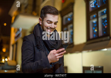 Homme habillé en style d'affaires sur la rue le soir se penche sur le smartphone et sourit. Banque D'Images