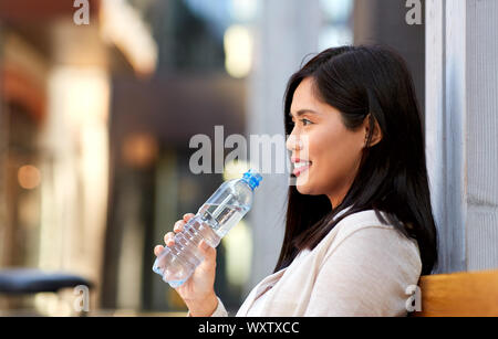 Mode de vie et de personnes concept - happy smiling young asian woman drinking water flom bouteille plastique assis sur un banc de la ville Banque D'Images