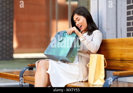 La vente, la consommation et les gens concept - étonné young asian woman with shopping bags assis sur un banc de jardin de ville Banque D'Images