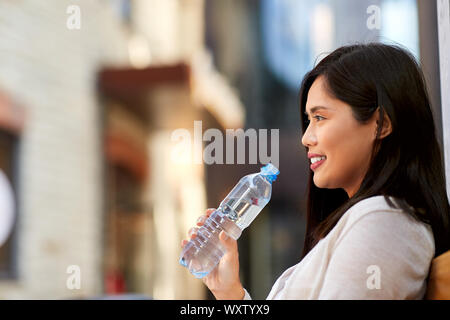 Mode de vie et de personnes concept - happy smiling young asian woman drinking water flom bouteille plastique assis sur un banc de la ville Banque D'Images