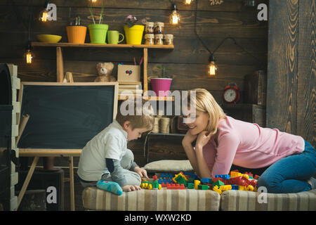 Vue de côté et cute boy smiling woman jouer avec des blocs de plastique. Maman se pencher en avant pour son fils assis sur le bord du canapé. Concept de la maternité. Banque D'Images