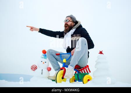 Lunettes pilote de Hipster et hat doigt pointé. Surpris man riding tricycle sur paysage enneigé. Noël et nouvel an plaisir. Maison de vacances célébration co Banque D'Images