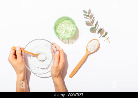 Les mains tenant bol de poudre d'argile cosmétique de faire masque de visage à côté de pot de gel d'aloès et brin d'eucalyptus sur fond blanc, télévision lay. Banque D'Images