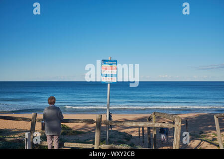 Une femme à côté d'une barrière en bois à l'entrée de Palm Beach avec un panneau d'avertissement sur une journée ensoleillée avec un ciel bleu clair, New South Wales, Australie Banque D'Images
