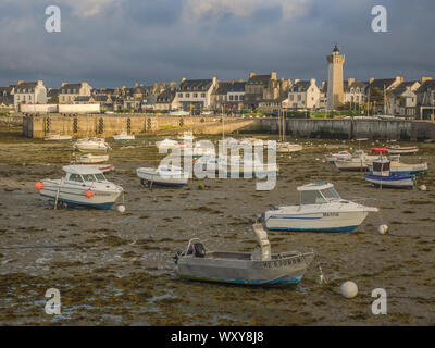 De nombreux petits bateaux assis dans la vase à marée basse dans le port de Roscoff, France, avec la ville et le phare éclairé par le soleil du soir. Banque D'Images