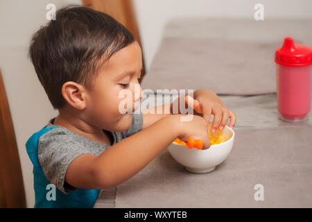 A cute toddler montre intérêt à manger un mac et fromage le déjeuner, et est titulaire d'cuillère pour auto-alimentation. Banque D'Images