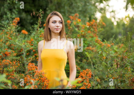 Cute femme en chemise jaune à côté de la rêverie smiling standing dans le jardin près de Pyracantha baies orange de buissons et d'arbres verts Banque D'Images
