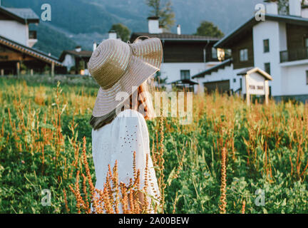 Splendida e romantica ragazza preteen nel cappello di paglia contro lo sfondo di belle case in montagna, rurale scena al tramonto. Vista da dietro, oro Foto Stock