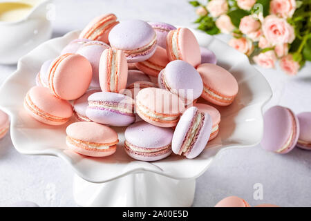 Rosa e lavanda francese macarons su una torta di porcellana stand con bouquet di rose e creamer in background, orizzontale vista da sopra Foto Stock