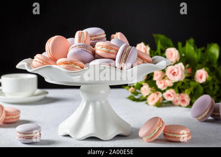 Close-up di macarons su una torta bianca stand. bel bouquet di rose e la tazza di caffè al fondo nero, spazio libero Foto Stock