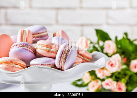 Close-up di macarons su un piatto bianco. bel bouquet di rose e un muro di mattoni in background Foto Stock