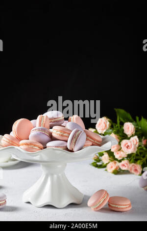 Rosa e lavanda macarons su una torta bianca stand con un bellissimo mazzo di rose a sfondo nero, vista verticale, close-up Foto Stock