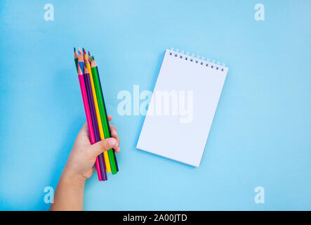 Bambino con le mani in mano azienda matite colorate e un notebook su sfondo blu con copyspace. Piatto stile di laici. Si torna a scuola concetto. Foto Stock