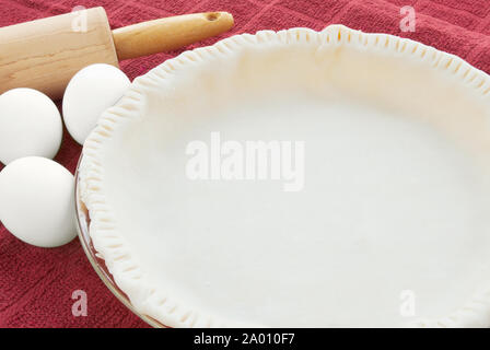 Vuoto pasta cruda base di pastafrolla per torta con bordi frastagliati in un grafico a torta di vetro piatto. Ci sono il bianco uova crude e un di legno mattarello in background. Foto Stock