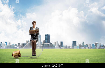 Giovane donna sorridente con apri libro seduti sulla grande lampadina. Bella ragazza in business suit su sfondo di paesaggio con il moderno centro citta', verde g Foto Stock