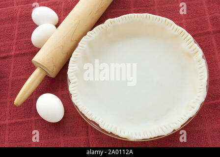 Vuoto pasta cruda base di pastafrolla per torta con bordi frastagliati in un grafico a torta di vetro piatto. Ci sono il bianco uova crude e un di legno mattarello in background. Foto Stock