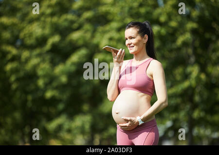 Giovane donna con capelli scuri in rosa abbigliamento sportivo di toccare la sua gravidanza ventre e la registrazione di un messaggio audio sul telefono cellulare all'aperto Foto Stock