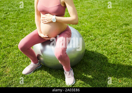 Close-up di giovane donna incinta in rosa abbigliamento sportivo seduti sulla sfera di fitness e toccando il suo ventre all'aperto Foto Stock