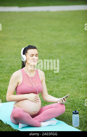 Giovane donna incinta nelle cuffie seduta sul tappetino di esercizio e godendo della buona musica durante il corso di formazione nel parco Foto Stock