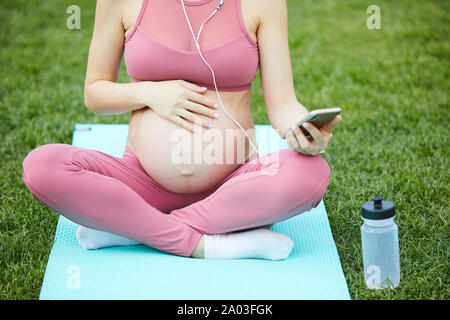 Close-up della donna incinta seduta sul tappetino di esercizio e ascoltare la musica sul suo telefono cellulare ha riposo dopo la formazione all'aperto Foto Stock